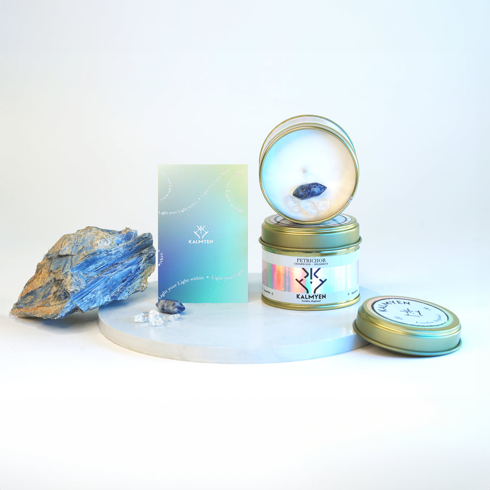 Image showing two Petrichore keepsake tin candles stacked on top of each other the top one is open showing the inside of the keepsake candle embedded with its Blue Spot Jasper mini crystal and clear quatz chips, the candles are diplayed on a blue disc with a raw crystal on the left, a gradient coloured scent card stands next to the raw crystal. 